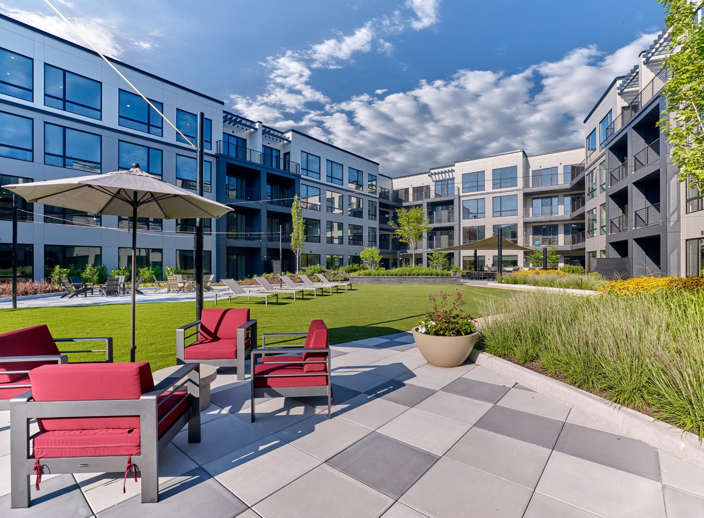 A sunny day at a modern apartment complex with red chairs and umbrellas on the patio.