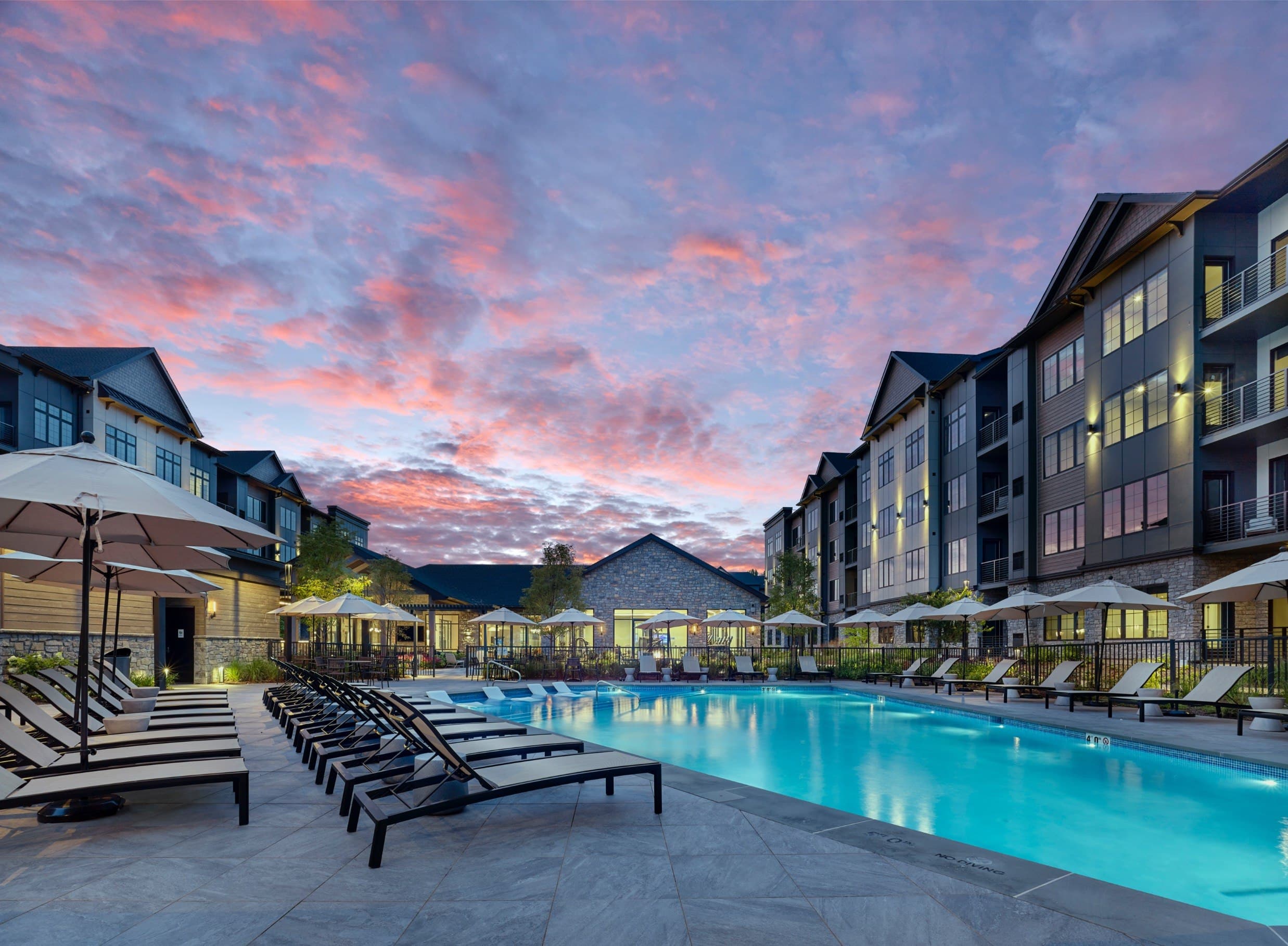 A beautiful outdoor swimming pool area with sun loungers and apartment buildings in the background during sunset.